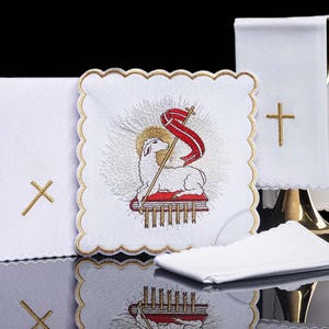 Può includere: Biancheria religiosa bianca con croci ricamate in oro e un pezzo quadrato centrale con un agnello che tiene uno stendardo rosso. La biancheria ha bordi smerlati ed è esposta su una superficie riflettente.