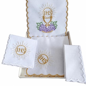 Può includere: Un set di biancheria liturgica bianca con bordi smerlati e finiture dorate. I pezzi sono ricamati con un simbolo IHS dorato circondato da raggi, un calice d'oro e uva viola con foglie verdi, il tutto presentato all'interno di una scatola.