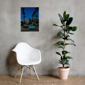 May include: A framed print depicts a nighttime street scene with a teal car driving towards city lights. Palm trees line the road under a dark blue sky. A white chair with wooden legs is in the foreground, and a potted plant is to the right.