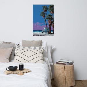 Puede incluir: Una impresión artística vertical que representa una escena de playa con palmeras, una torre de salvavidas y un cielo vibrante. La obra de arte presenta un cielo azul con una luna, nubes rosas y un océano turquesa. La impresión se muestra en una pared blanca sobre una cama.
