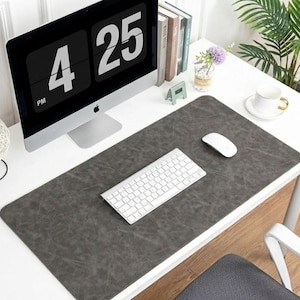 Pode incluir: Um tapete de mesa retangular cinza escuro com uma superfície texturizada. Um teclado e um rato brancos estão sobre o tapete, juntamente com um monitor de computador que mostra a hora 16:25. A mesa é branca, com uma chávena e livros visíveis.