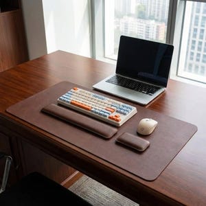 Puede incluir: Un tapete de escritorio de cuero marrón con un reposamuñecas y un mousepad a juego. Un teclado mecánico crema con teclas azules y naranjas se encuentra sobre el tapete, junto con una computadora portátil y un mouse inalámbrico. El escritorio es de madera oscura.