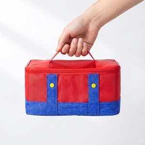 May include: Red and blue insulated lunch bag with a top handle. The bag has a red top and a blue bottom section with two blue vertical straps and yellow circular accents. The bag is held by a hand.