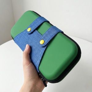 May include: Green rectangular case with a blue denim strap secured by two yellow buttons. The case has a black zipper closure. The case is held in a person's hand.