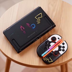 May include: Black handheld gaming console with a matching controller. The console features neon graphics and the text "ANNIVERSARY XV". The controller has black, white, and orange accents.