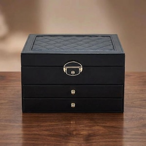 Może przedstawiać: Czarna szkatułka na biżuterię z pikowaną górą i dwiema szufladami. Pudełko ma złote zapięcie i gałki szuflad. Szkatułka na biżuterię wykonana jest z gładkiego materiału i stoi na drewnianej powierzchni.