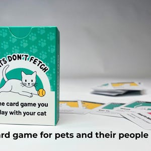 May include: A teal and white card game box titled Cats Don't Fetch with a white cat illustration and a yellow ball. The box text reads The card game you play with your cat. Several playing cards with cat graphics are scattered on the surface.