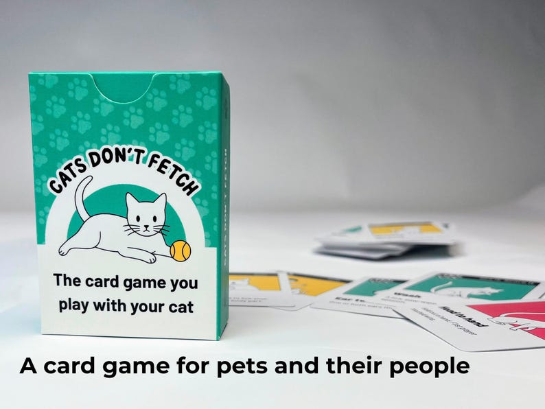 May include: A teal and white card game box titled Cats Don't Fetch with a white cat illustration and a yellow ball. The box text reads The card game you play with your cat. Several playing cards with cat graphics are scattered on the surface.