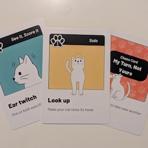 May include: Three rectangular game cards featuring white cat illustrations. The left card is teal with the text See It, Score It and Ear twitch. The center card is yellow with the text Solo and Look up. The right card is orange with Chaos Card text.