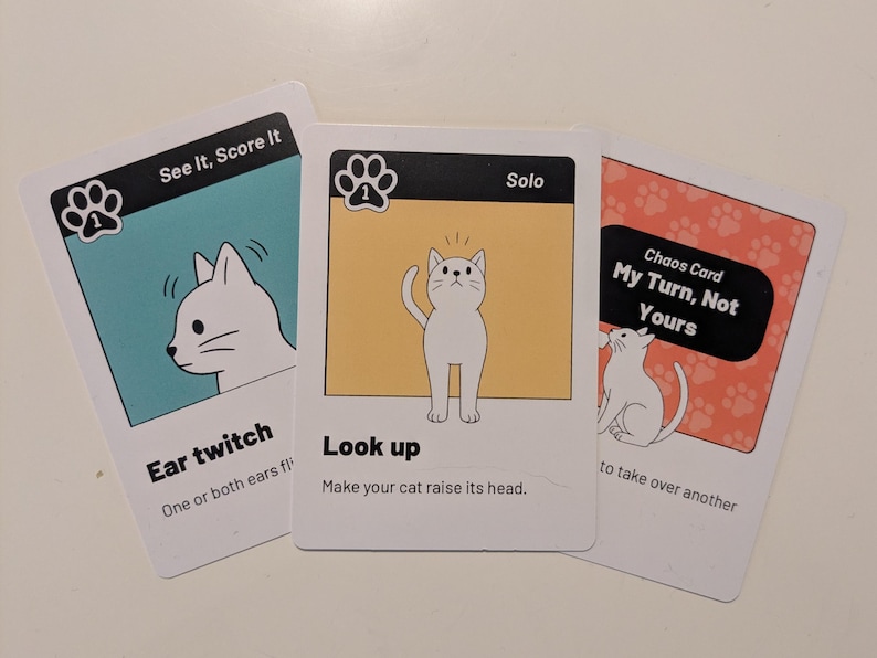 May include: Three rectangular game cards featuring white cat illustrations. The left card is teal with the text See It, Score It and Ear twitch. The center card is yellow with the text Solo and Look up. The right card is orange with Chaos Card text.