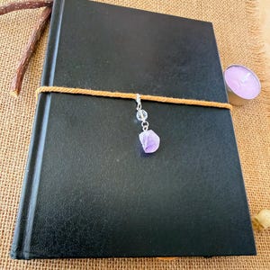 Può includere: Un diario con copertina rigida nera avvolto da un cordoncino di lana color marrone chiaro. Un ciondolo color argento pende dal cordoncino, caratterizzato da una piccola perla trasparente sfaccettata e da una pietra ametista viola grezza.