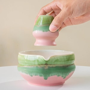 Puede incluir: Un juego de cuenco y batidor de matcha de cerámica. El cuenco es rosa y verde con un interior blanco. El batidor es verde y rosa. El juego está diseñado para preparar té matcha.
