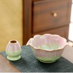 May include: A ceramic matcha bowl and whisk holder in a soft pink and green glaze. The bowl has a lotus flower shape with a scalloped rim. The whisk holder is small and egg-shaped. Both items are on a dark grey surface.
