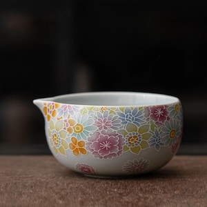 Puede incluir: Una jarra de té de cerámica con un estampado floral. La jarra es de color gris claro con un diseño colorido de flores en tonos rosa, amarillo, naranja y azul. Tiene un pico.