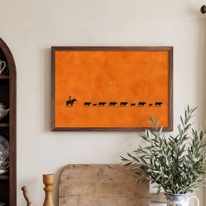 May include: A rectangular wall art print featuring a solid orange background with a black silhouette of a person riding a horse followed by a line of cattle. The print is enclosed in a thin, dark brown wooden frame and mounted on a wall.