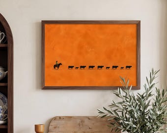 May include: A rectangular wall art print featuring a solid orange background with a black silhouette of a person riding a horse followed by a line of cattle. The print is enclosed in a thin, dark brown wooden frame and mounted on a wall.