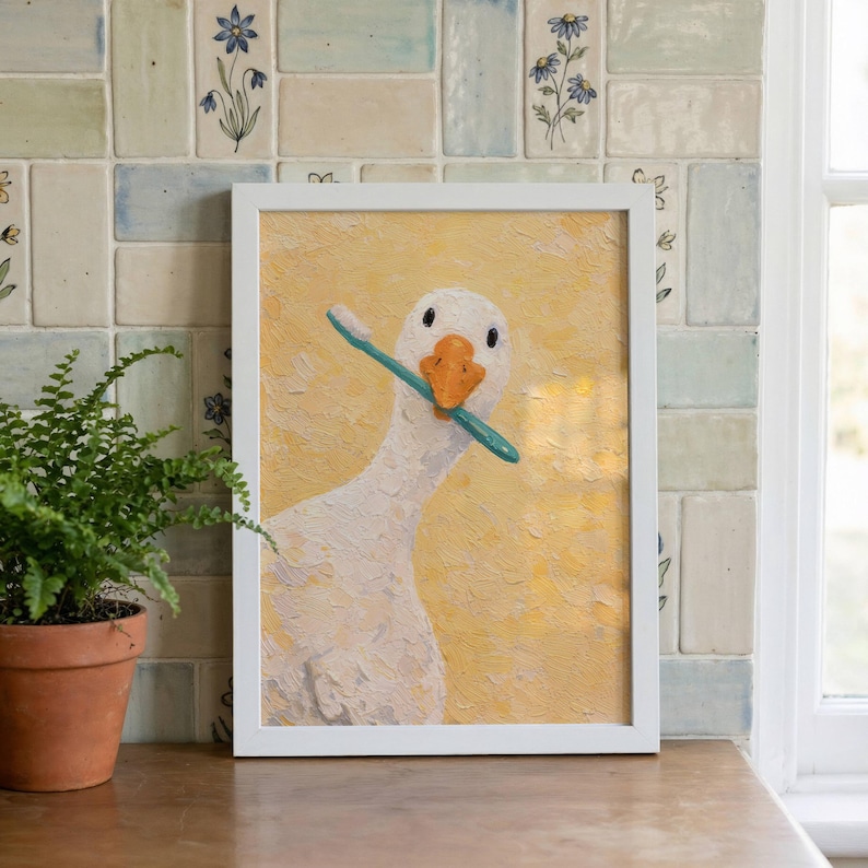 May include: A framed art print depicting a goose with a teal toothbrush in its beak. The goose is painted white with an orange beak, set against a textured yellow background. The artwork is presented in a white frame.