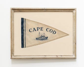 May include: A framed print featuring a triangular pennant flag in cream and navy blue. The flag displays the text CAPE COD in navy blue lettering above a detailed illustration of a fishing boat. The artwork is enclosed in a light wood frame.