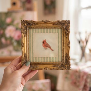 May include: A small, ornate gold picture frame with a detailed border. Inside, a watercolor painting of a red cardinal bird is set against a striped background. The frame is held up by a hand, showcasing the artwork.