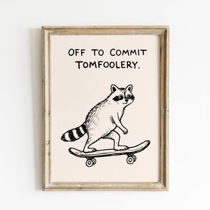 May include: A framed wall art print featuring a black and white line drawing of a raccoon riding a skateboard. Above the raccoon, the text reads OFF TO COMMIT TOMFOOLERY. The print is enclosed in a light-colored wooden frame with a visible grain.