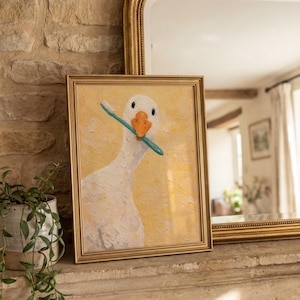 May include: A framed picture of a white goose with a blue toothbrush in its beak, set against a yellow backdrop. The artwork is in a gold frame. The picture is displayed on a stone surface.
