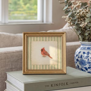 May include: A framed print featuring a watercolor painting of a red cardinal bird. The artwork is set against a striped background and framed in gold. The frame rests on a stack of books, with a vase of flowers in the background.