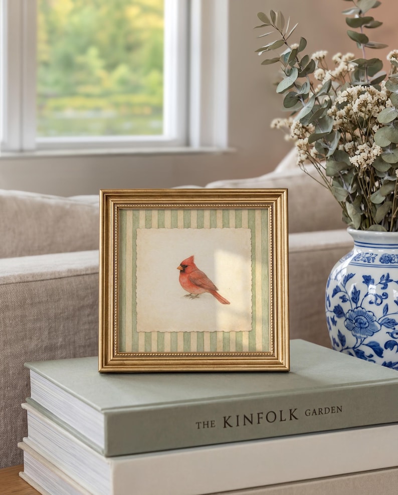 May include: A framed print featuring a watercolor painting of a red cardinal bird. The artwork is set against a striped background and framed in gold. The frame rests on a stack of books, with a vase of flowers in the background.