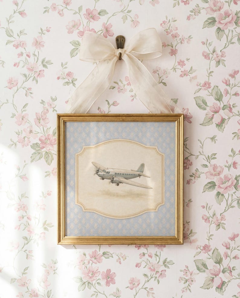 May include: A framed print of a vintage airplane, set against a light blue background with a decorative border. The gold-colored frame is suspended by a cream-colored bow on a floral wallpaper background. The print is a charming piece of wall decor.