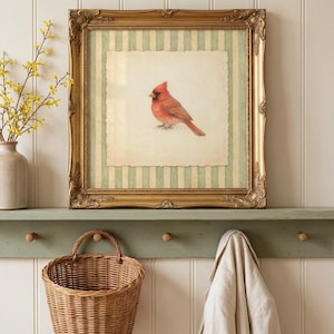 May include: A framed print featuring a red cardinal bird. The print is set against a cream-colored background with green and white stripes, within a gold ornate frame. A woven basket and a coat hang below.