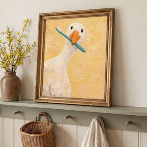 May include: A framed painting of a white goose with an orange beak, holding a blue toothbrush. The artwork is set against a textured yellow background and is enclosed in a gold-coloured frame. The painting is a humorous piece.