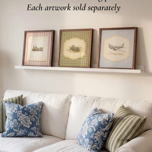 May include: Three framed artworks with vintage transportation illustrations: a train, a car, and an airplane. The frames are dark wood, and the prints have a soft, aged appearance. The artworks are displayed above a white couch with blue floral and green striped pillows.