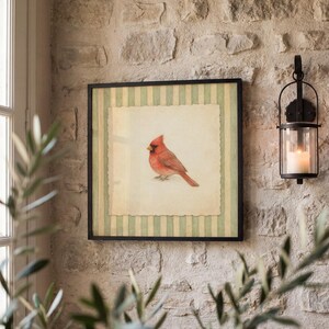 May include: A framed artwork featuring a red cardinal bird. The artwork has a cream-colored background with a green and white striped border. The frame is black, and the artwork is hanging on a textured stone wall. A black lantern with a lit candle is to the right.