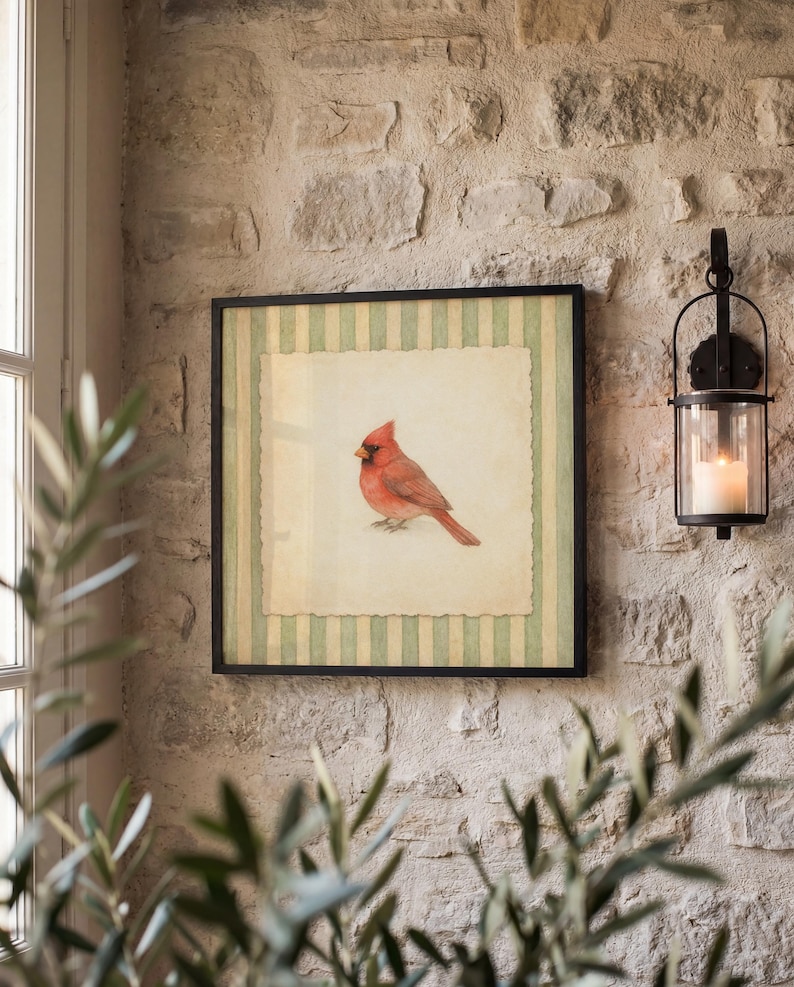 May include: A framed artwork featuring a red cardinal bird. The artwork has a cream-colored background with a green and white striped border. The frame is black, and the artwork is hanging on a textured stone wall. A black lantern with a lit candle is to the right.