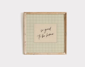 May include: A square wooden frame containing a beige card with the black cursive text so good to be home, centered over a light sage green and white gingham patterned background. The frame has a natural wood finish with visible grain texture.