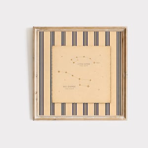 May include: A square framed print featuring a beige background with vertical navy blue stripes. Centered is a cream-colored paper with deckled edges displaying constellation diagrams labeled Little Dipper and Big Dipper in dark brown text.