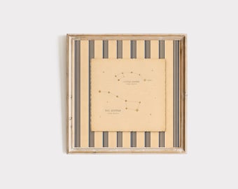May include: A square framed print featuring a beige background with vertical navy blue stripes. Centered is a cream-colored paper with deckled edges displaying constellation diagrams labeled Little Dipper and Big Dipper in dark brown text.