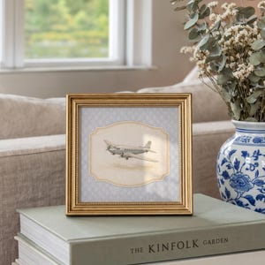 May include: A gold-framed picture featuring a vintage airplane illustration. The artwork is set against a light blue patterned background. The frame rests on a stack of books, with a vase of flowers in the background. The book on top is titled "THE KINFOLK GARDEN."