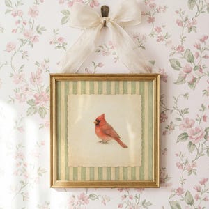 May include: A framed print featuring a red cardinal bird. The artwork is set within a gold frame with a green and cream striped border. A cream-colored bow hangs above the frame, against a floral wallpaper background.