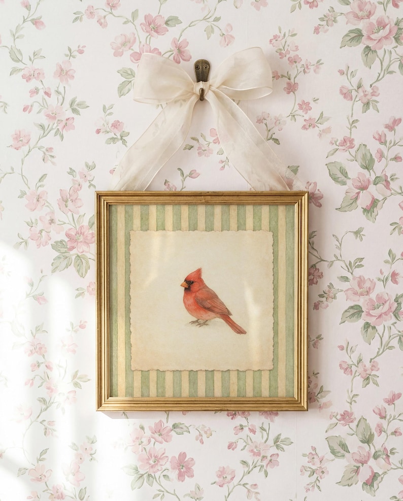 May include: A framed print featuring a red cardinal bird. The artwork is set within a gold frame with a green and cream striped border. A cream-colored bow hangs above the frame, against a floral wallpaper background.