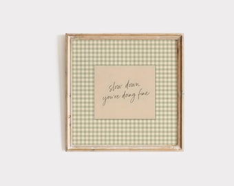 May include: A square wooden frame containing a light beige card with the black cursive text 'slow down you're doing fine' centered over a background featuring a light green and white gingham check pattern within the frame's border.