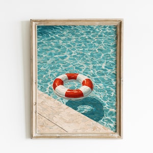 Op de afbeelding: Ingelijste kunstdruk van een zwembad met een rood-witte reddingsboei die in het turquoise water drijft. De lijst is lichtbruin.