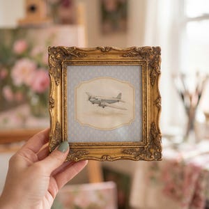 May include: A small, ornate gold picture frame holding a vintage-style print of an airplane. The print is set against a light blue patterned background. The frame is held by a hand with a painted fingernail.