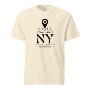 May include: Cream-colored t-shirt featuring a black graphic of a map pin over a map outline, with "NY" below. The coordinates 40.7128 N / 74.0060 W and "Spawn Point" are printed below the letters. The shirt has a crew neck and short sleeves.