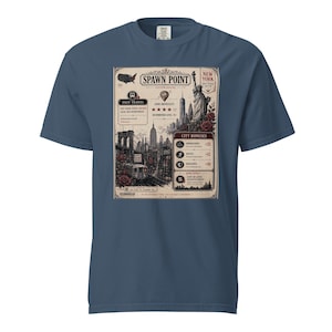 May include: A blue t-shirt featuring a vintage-style graphic of New York City. The design includes the Statue of Liberty, a cityscape, and the words "Spawn Point" and "New York". The shirt has a crew neck and short sleeves.