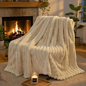 May include: A cream-colored, plush throw blanket draped over a chair. The blanket has a ribbed texture and a soft, inviting appearance. A lit candle in a glass jar sits on a wooden tray in the foreground. The word "kyroo" is printed on the candle.