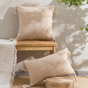 Peut inclure: Deux coussins beiges tissés avec des glands décoratifs. Un coussin carré repose sur un banc en bois, l'autre coussin rectangulaire est sur un tabouret circulaire tissé. Les coussins ont un motif texturé et sont de couleur neutre.