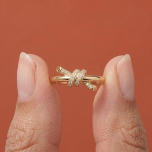 May include: A gold-colored ring with a knot design, encrusted with small, clear gemstones. The ring is held between two fingers against a solid orange-red background. The ring's design features a knot shape.
