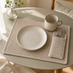 Puede incluir: Un mantel individual rectangular de ganchillo en color beige con un bolsillo a juego para los cubiertos. El conjunto incluye un plato redondo de cerámica beige, una taza de cerámica beige, un tenedor de plata y un cuchillo de plata.