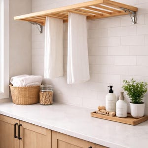 Könnte beinhalten: Ein Handtuchhalter aus hellem Holz mit zwei weißen Handtüchern, die daran hängen, an einer weiß gefliesten Wand befestigt. Ein Weidenkorb mit gefalteten weißen Handtüchern, ein Glas und ein Holztablett mit Seifenflaschen und einer Pflanze befinden sich auf der Arbeitsplatte.