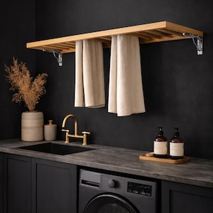 Könnte beinhalten: Ein moderner Waschraum mit schwarzer Arbeitsplatte, Schränken und einer Waschmaschine. Ein Handtuchhalter aus Holz hält zwei beige Handtücher. Ein goldener Wasserhahn und ein Waschbecken sind sichtbar, ebenso wie dekorative Flaschen und eine Vase.